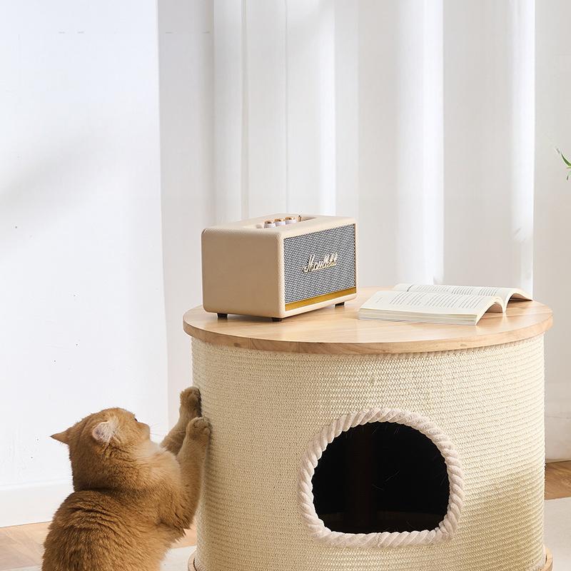 A round big cat condo with a woven cream body, a light wood top, and a circular opening on the front, shown in a bright room. An orange tabby cat is climbing on the left side, and a small cream speaker and an open book sit on top.