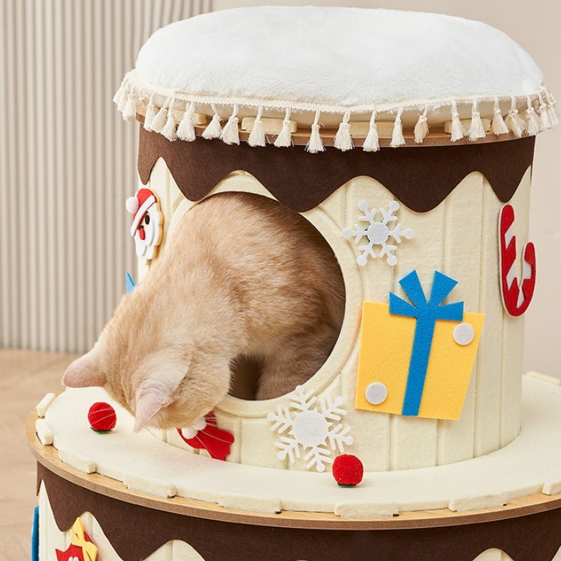 A light brown cat peers into the circular opening of a decorative cat condo designed to resemble a frosted dessert or gift box, with cream-colored panels, white tassel trim, brown icing-like accents, and festive felt decorations, including a blue gift box, white snowflakes, and red candy details.