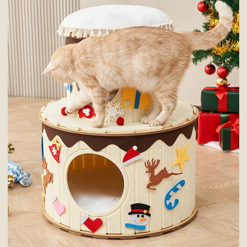 A large orange cat stands on a round, Christmas-themed cat cave in a decorated living room. The cat condo is cream-colored with brown trim, decorated with felt snowmen, reindeer, a Santa hat, candy canes, and stars, and has a small white-tufted top section.