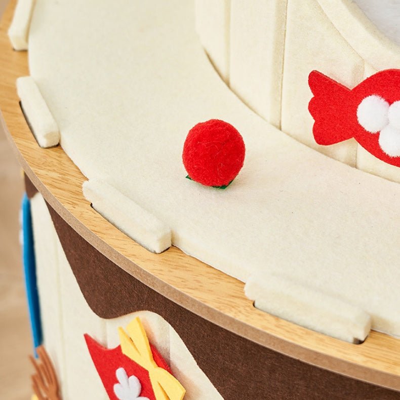Close-up of a wooden cat condo with cream-colored felt sails, brown hull details, and small red felt fish accents, with a small red ball on the deck.