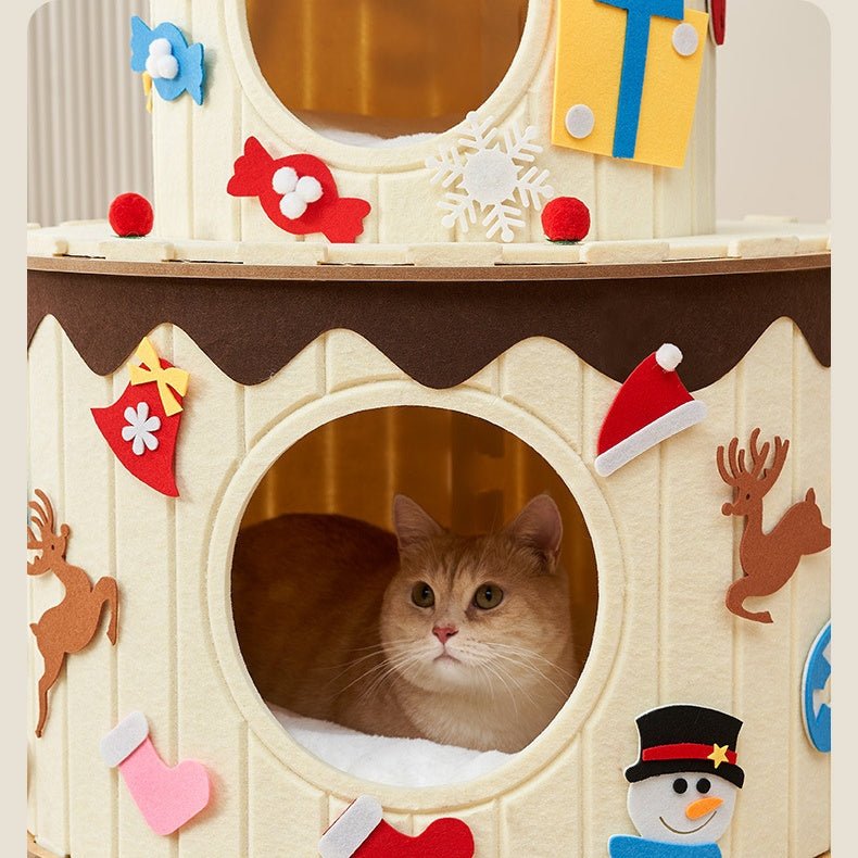 A cream-colored cat tree with a round window opening showing a fluffy orange cat inside. The birthday cake cat tree is decorated with festive Christmas felt cutouts, including a reindeer, stocking, snowman, candy, and snowflake, with a small yellow gift box hanging at the top.
