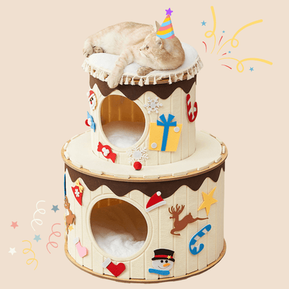 A two-tiered cat condo styled like a decorated cake, with a light-colored cat lying on top. The tree has cream-colored panels, dark brown frosting-like trim, and festive holiday decorations including Santa hats, a reindeer, a snowman, and gift boxes.