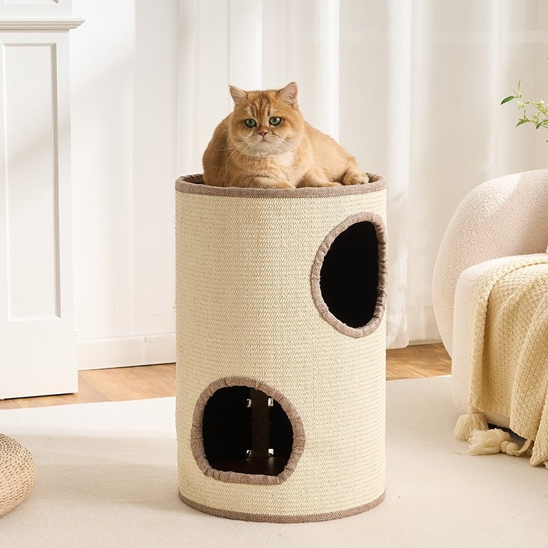 A ginger cat sits inside a tall cylindrical corner cat condo with two circular openings, placed in a bright, neutral-toned room with curtains, a chair, and a blanket visible.