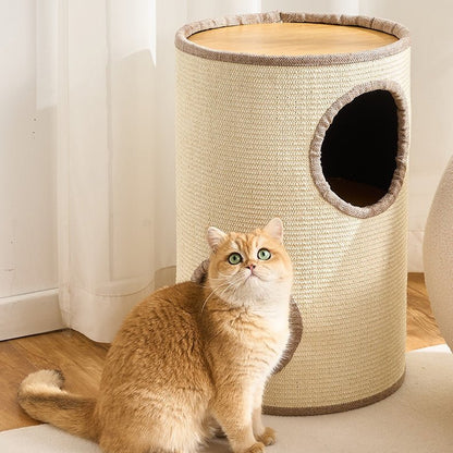 A ginger cat sits in front of a cylindrical beige cat condo with a woven texture, a round wooden top, and a dark cat hole on a light wood floor near white curtains.