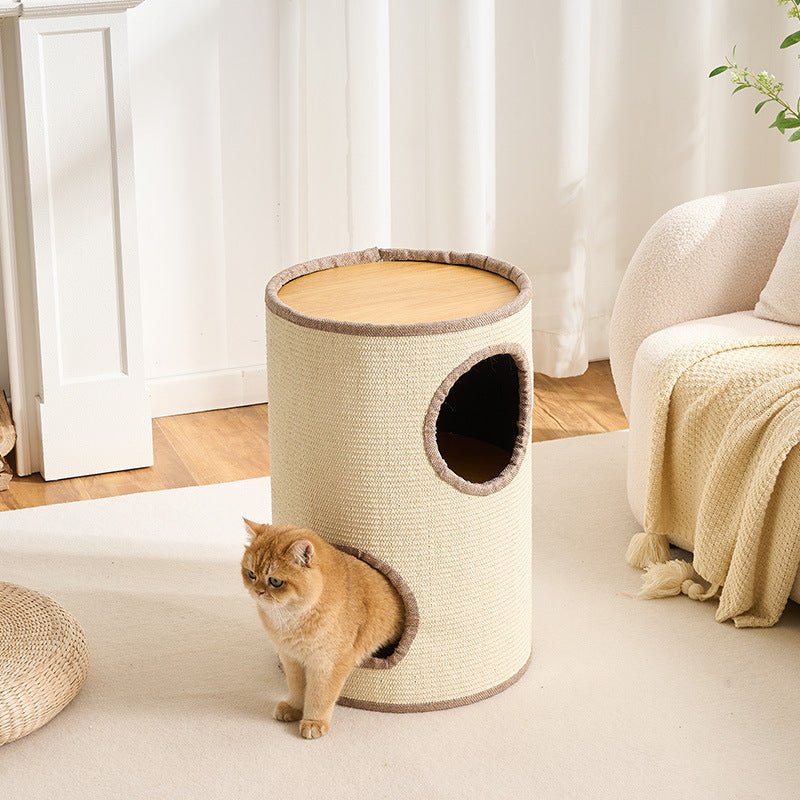 A cylindrical cat tower with a woven beige body, light wood top, and two openings, shown in a bright living room with a ginger cat standing near the base.
