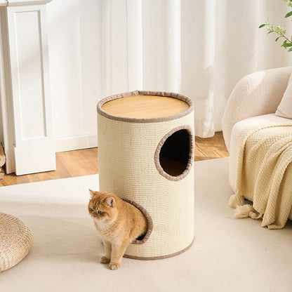 A cylindrical cat tower with a woven beige body, light wood top, and two openings, shown in a bright living room with a ginger cat standing near the base.