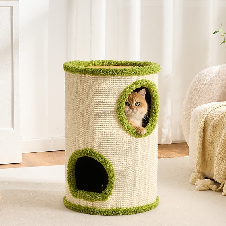 A large cat tower with a natural sisal-wrapped body and green faux-fur trim, featuring a top platform, a lower dark entry hole, and a circular window showing a ginger cat peeking out. The tower is set in a bright, modern room with white curtains and a neutral sofa in the background.