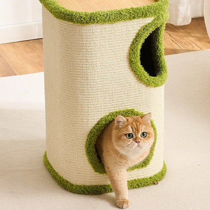 A golden cat stands in the front opening of a cylindrical cat tower with a cream woven body and bright green trim, photographed indoors on a light rug with wood flooring in the background.