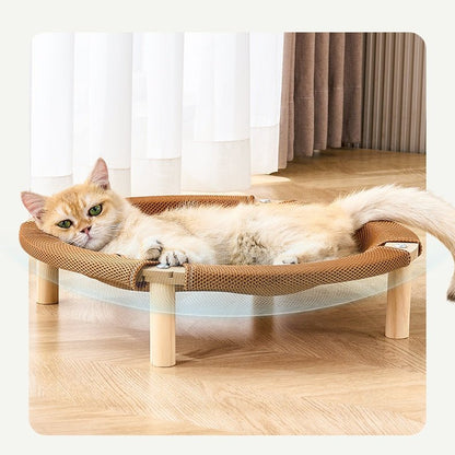 A cream-colored cat lies stretched out on a round woven cat bed with wooden legs in a bright room with light wood floors and curtains.