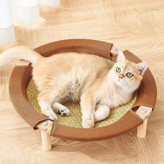 A fluffy cream-colored fat cat lies on a round woven pet bed with a brown mesh outer rim and light wood legs, placed on a light wood floor.