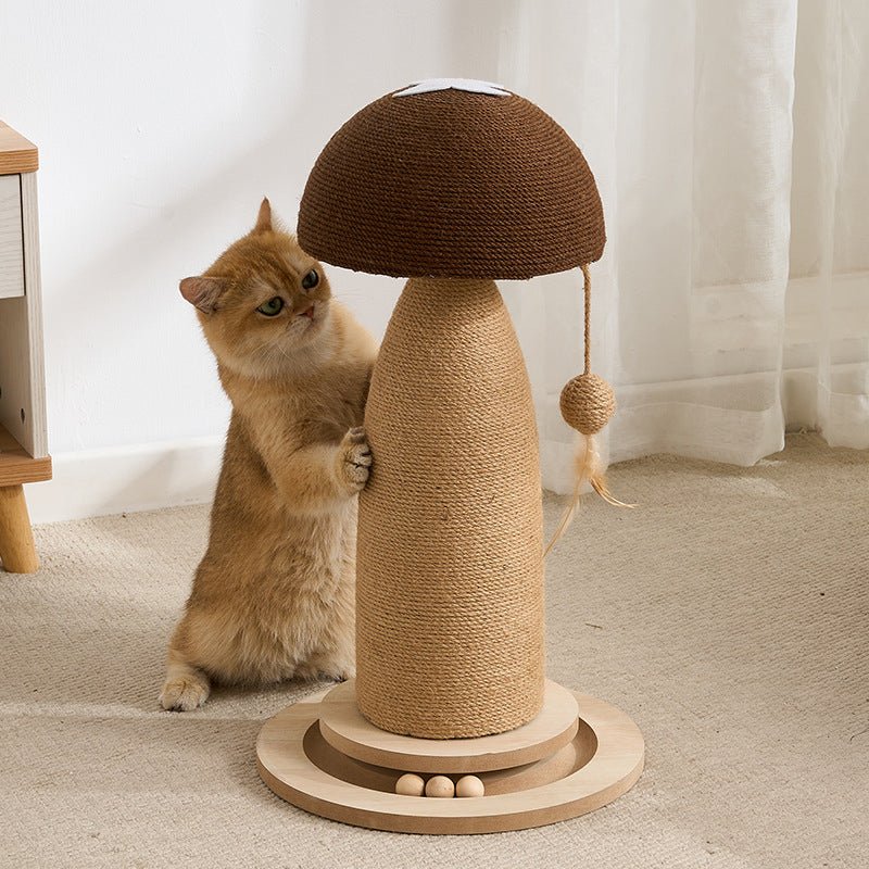 A fluffy golden cat stands on its hind legs beside a mushroom-shaped cat toy in a bright indoor room. The toy has a brown woven cap, a natural jute-wrapped body, a dangling sisal ball, and a round wooden base with small wooden beads.