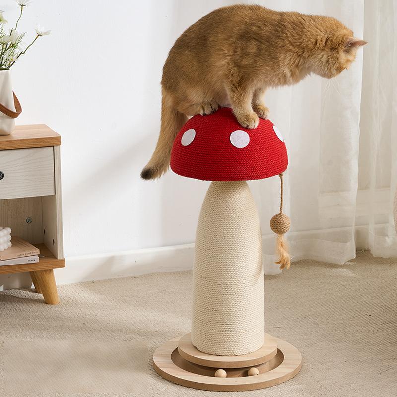 A ginger cat sits on a mushroom-shaped cat scratching post in a bright room. The post has a red cap with white polka dots, a beige wrapped body, and a round wooden base.