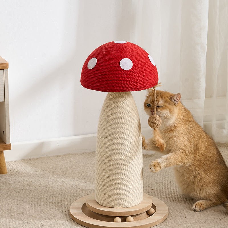 A ginger cat sits beside a mushroom-shaped cat scratching post in a bright room with white curtains and a light carpet. The red post has a red woven cap with white polka dots, a beige wrapped post, and a round wooden base.