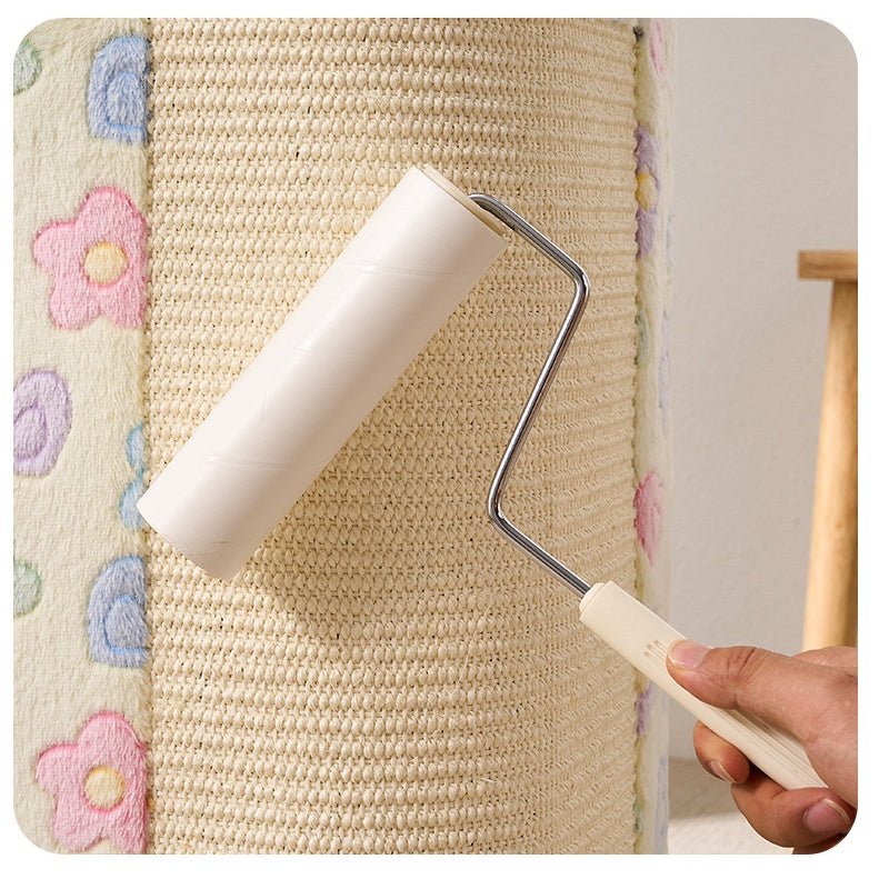 A hand holding a small paint roller with an off-white handle and a clear roller sleeve, applying cream-colored paint to a tall woven scratching post or house. The post features a vertical textured weave and a vertical panel on the left with pastel embroidered flowers in pink, blue, and purple on a beige background.