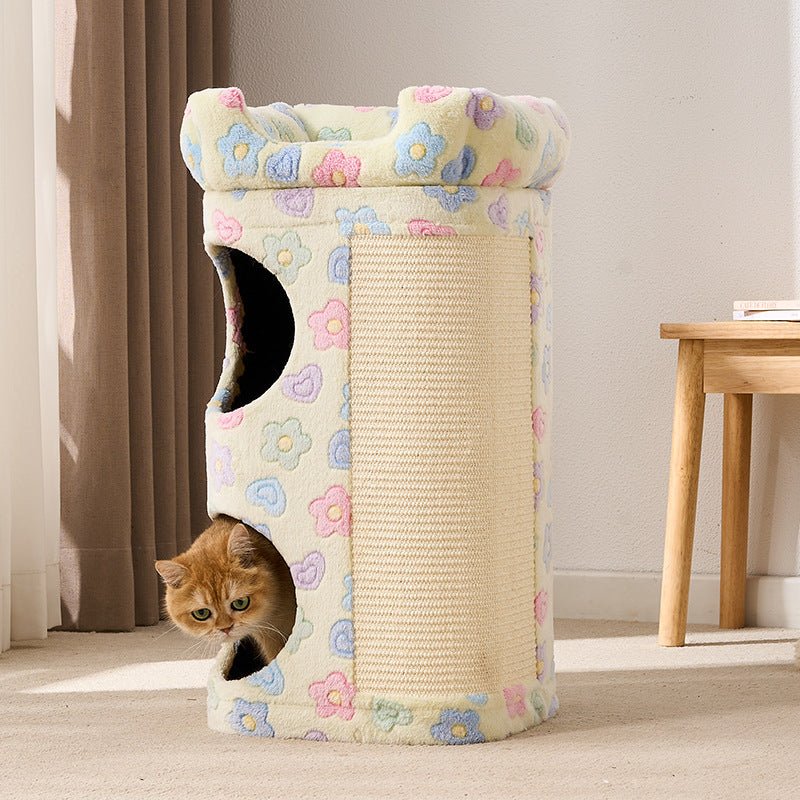 A cylindrical cat tower with a cream-colored floral fabric exterior, a textured scratching panel on the side, and two openings, one of which is shown with an orange cat peeking out. The setting is a cozy indoor room with curtains and a wooden table in the background.
