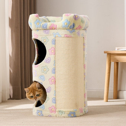 A cylindrical cat tower with a cream-colored floral fabric exterior, a textured scratching panel on the side, and two openings, one of which is shown with an orange cat peeking out. The setting is a cozy indoor room with curtains and a wooden table in the background.
