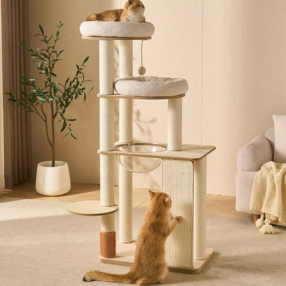 A large multi-level cat tree in light wood and cream fabric stands in a softly lit living room. Two orange tabby cats are present: one resting on the top platform and one scratching at the lower level.